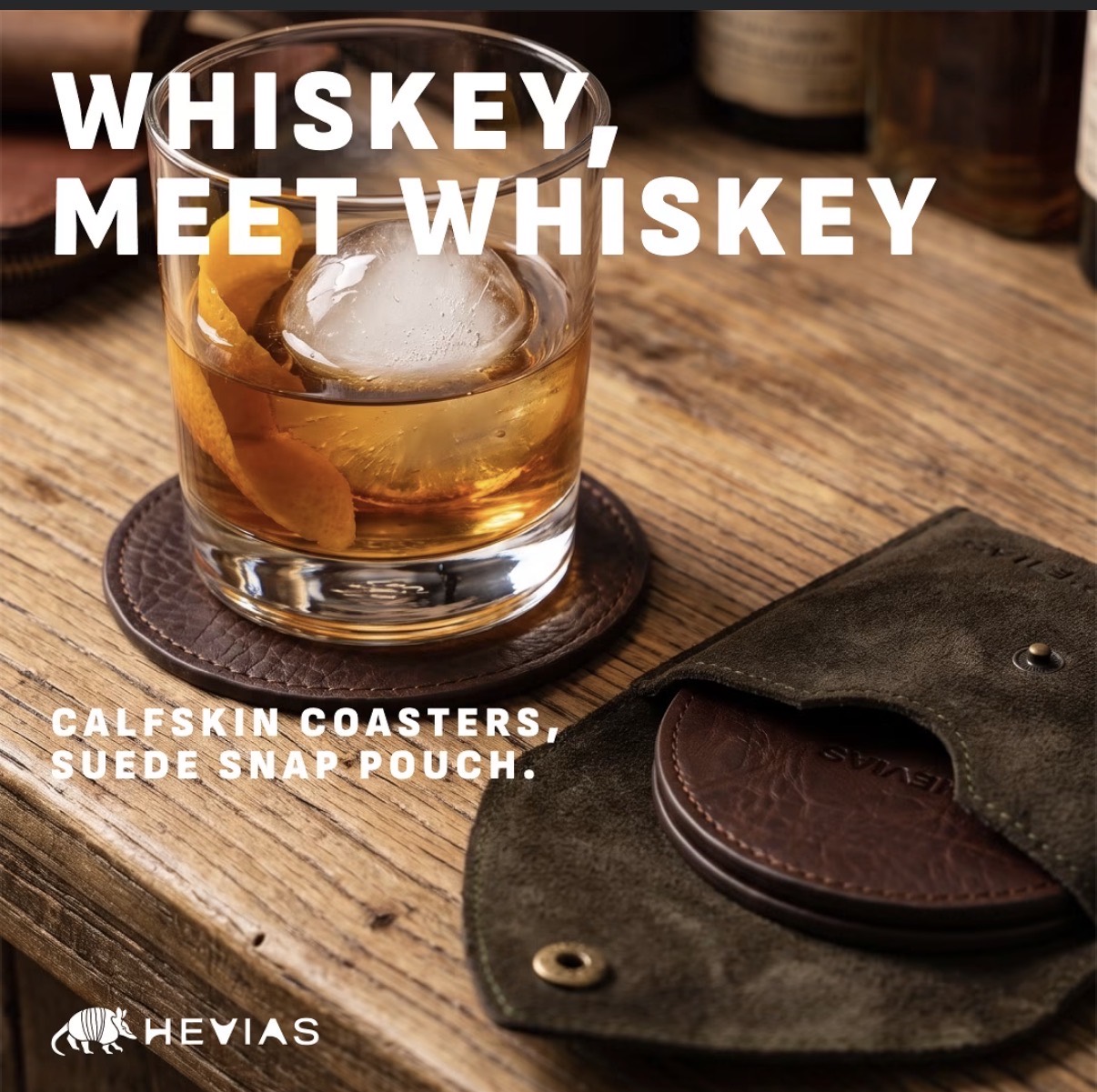 Hevias promotional artwork showing a whiskey cocktail on a brown leather coaster beside a suede snap pouch