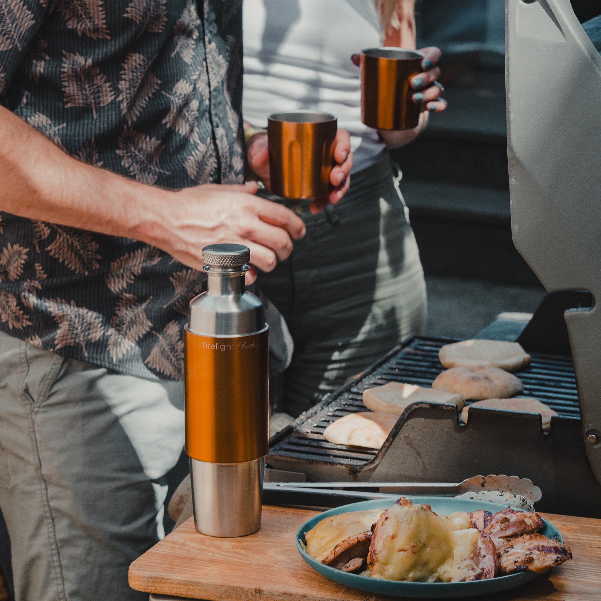 Copper Firelight Flask beside a grill with two people holding its matching tumblers