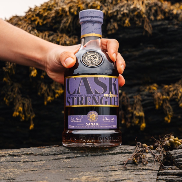 A hand holding the purple-labelled Sanaig Cask Strength 2026 bottle against coastal rocks
